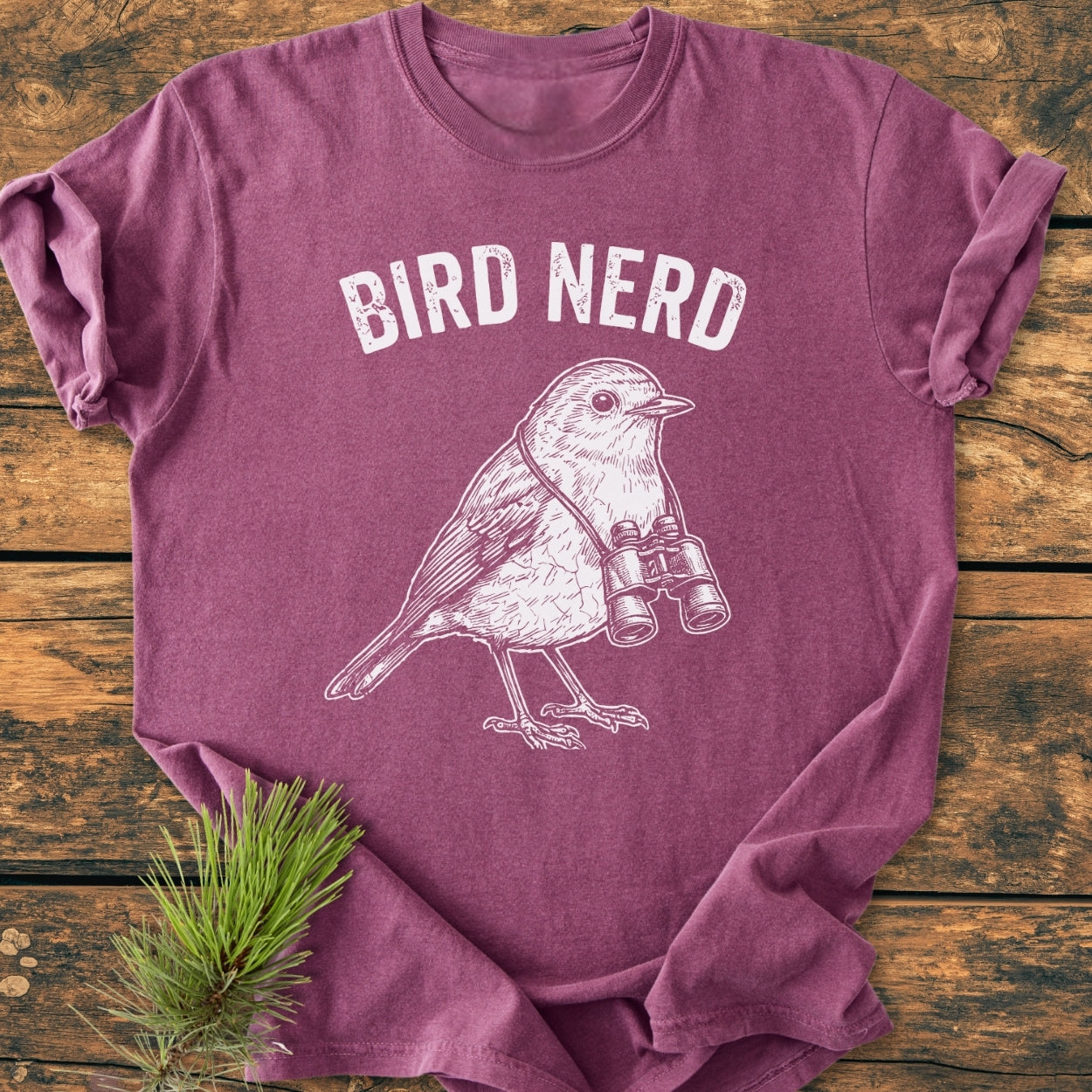 The Bird Nerd - Vintage Wash Tee features a heather-purple design with BIRD NERD and a songbird illustration. It's laid out on wood with pine sprigs, showcasing its relaxed, durable construction.