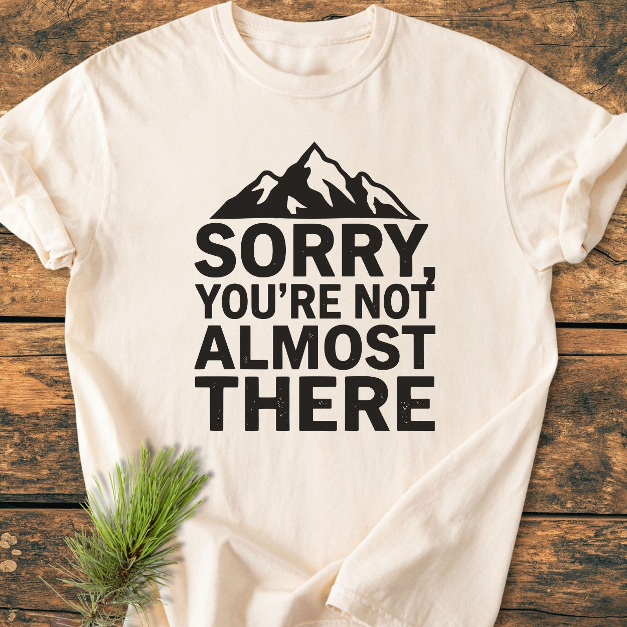 Sorry You're Not Almost There - Vintage Wash Tee features a light-cream hue with a mountain silhouette and bold text. A spruce twig beside it enhances the outdoor aesthetic.