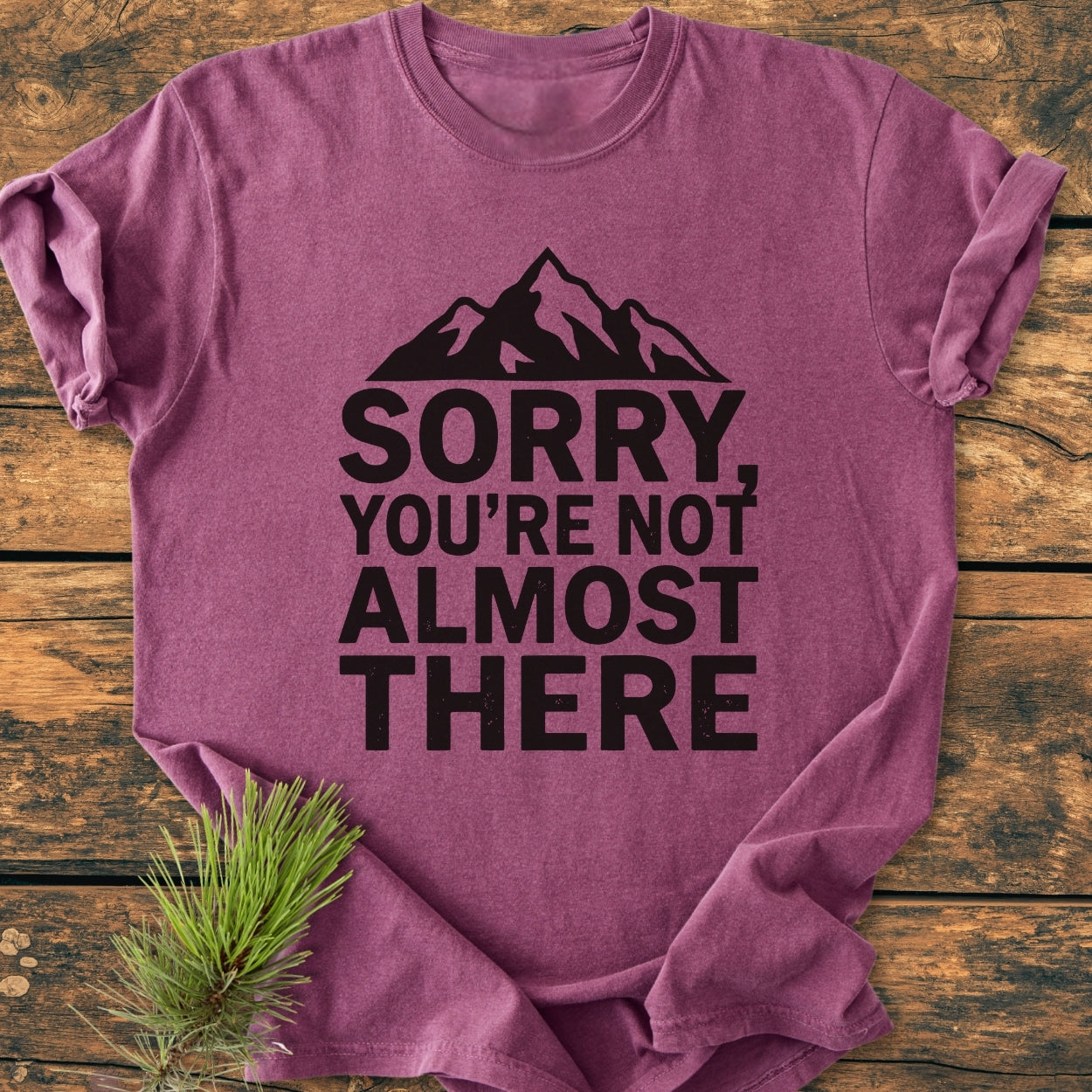 Folded Sorry You're Not Almost There - Vintage Wash Tee on wood surface, featuring a stylized mountain graphic and bold text, with a pine sprig beside it, highlighting its relaxed fit and durability.