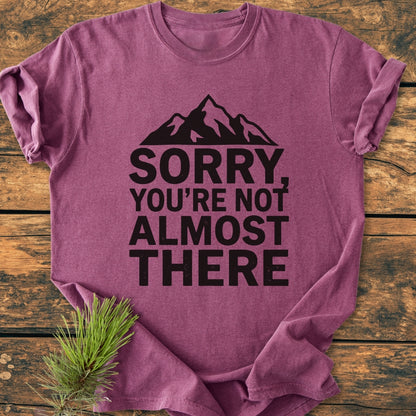 Folded Sorry You're Not Almost There - Vintage Wash Tee on wood surface, featuring a stylized mountain graphic and bold text, with a pine sprig beside it, highlighting its relaxed fit and durability.
