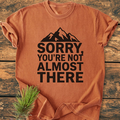 Rust-orange Sorry You're Not Almost There - Vintage Wash Tee with a black mountain silhouette. Laid flat on wood, styled with a pine sprig, showcasing cozy, durable, garment-dyed, ring-spun cotton.