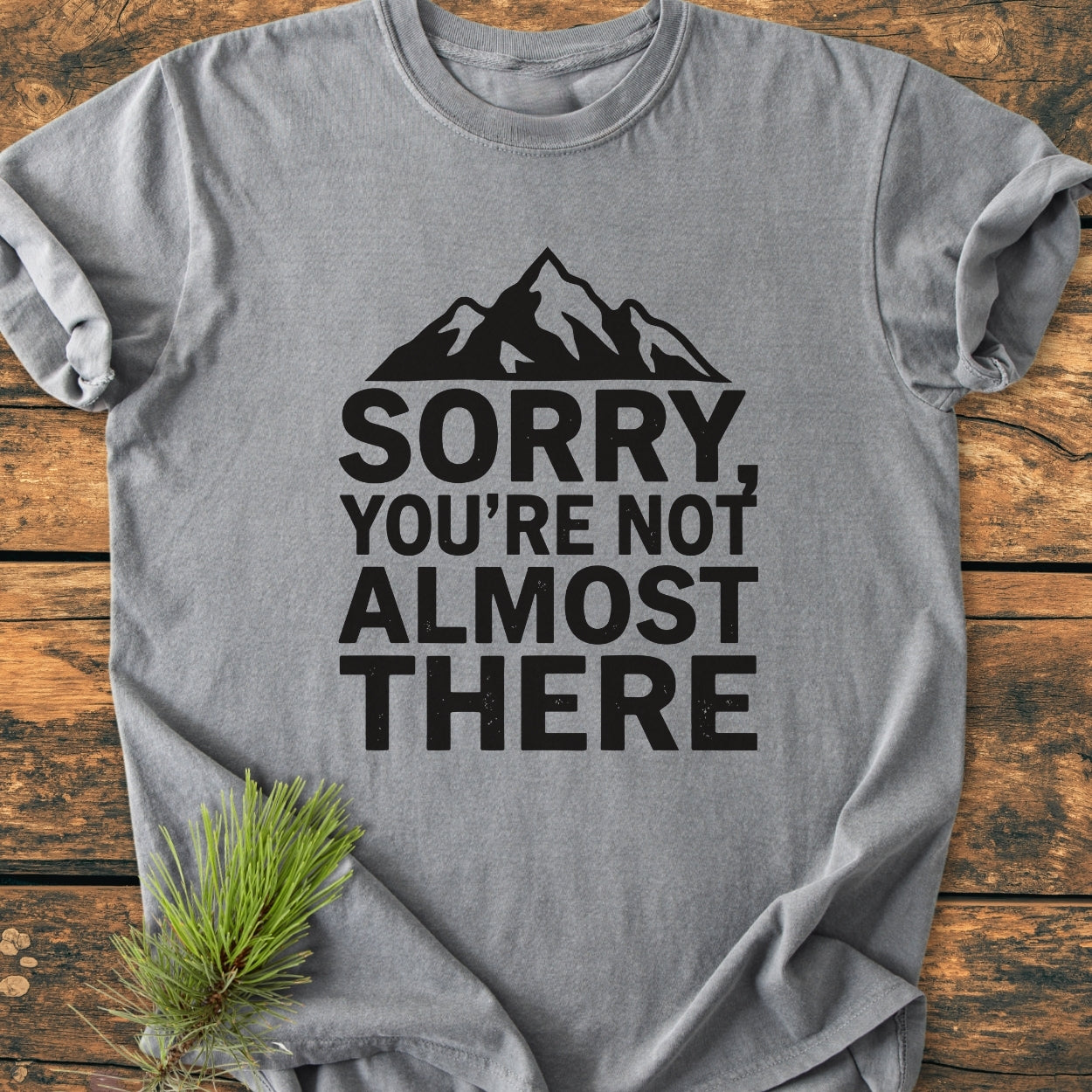 Sorry You're Not Almost There - Vintage Wash Tee featuring a black mountain graphic and bold text on a gray backdrop, laid on a wooden surface, highlighting its relaxed, garment-dyed fabric.