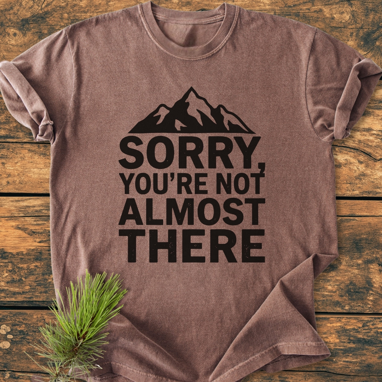 Sorry You're Not Almost There - Vintage Wash Tee, brown with a black mountain silhouette and text, laid on wood. Rolled sleeves and pine sprig evoke an outdoorsy vibe, enhancing its casual appeal.