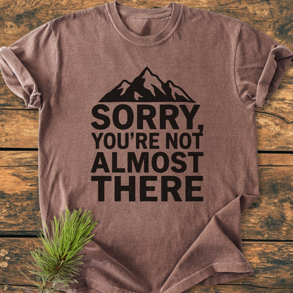 Sorry You're Not Almost There - Vintage Wash Tee, brown with a black mountain silhouette and text, laid on wood. Rolled sleeves and pine sprig evoke an outdoorsy vibe, enhancing its casual appeal.