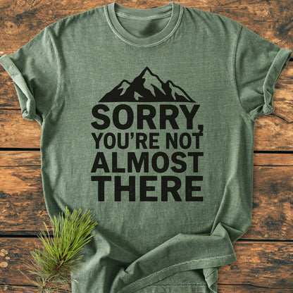 The Sorry You're Not Almost There - Vintage Wash Tee features a mountain silhouette and bold text on a folded green T-shirt, displayed on a wooden surface with a pine sprig beside it.