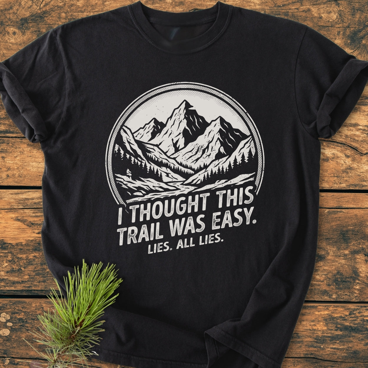 Lies, All Lies - Vintage Wash Tee featuring a mountain and forest graphic, laid on wood. The black Comfort Colors 1717 T-shirt is soft, durable, and ideal for casual wear.