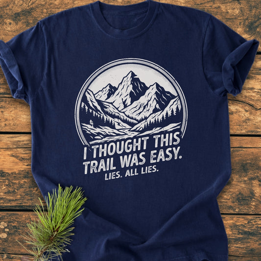 Lies, All Lies - Vintage Wash Tee features a hand-drawn mountain scene with humorous text, styled with a pine sprig on a wooden background, showcasing the tee’s relaxed fit and durable design.