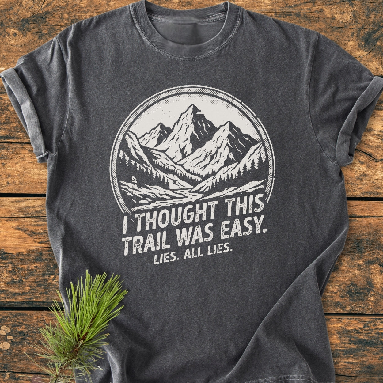 The Lies, All Lies - Vintage Wash Tee features a white mountain and tree graphic with humorous text on a charcoal-gray background, laid on a wooden surface with a pine sprig beside it.