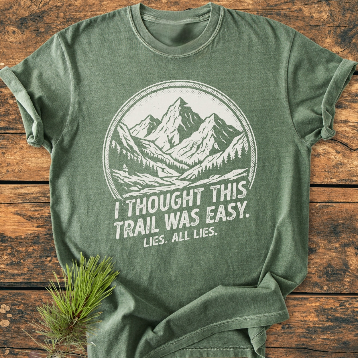 Lies, All Lies - Vintage Wash Tee: Heather-green short-sleeve T-shirt featuring a white circular mountain illustration with trees and trail, paired with humorous slogan, laid flat on wooden surface.