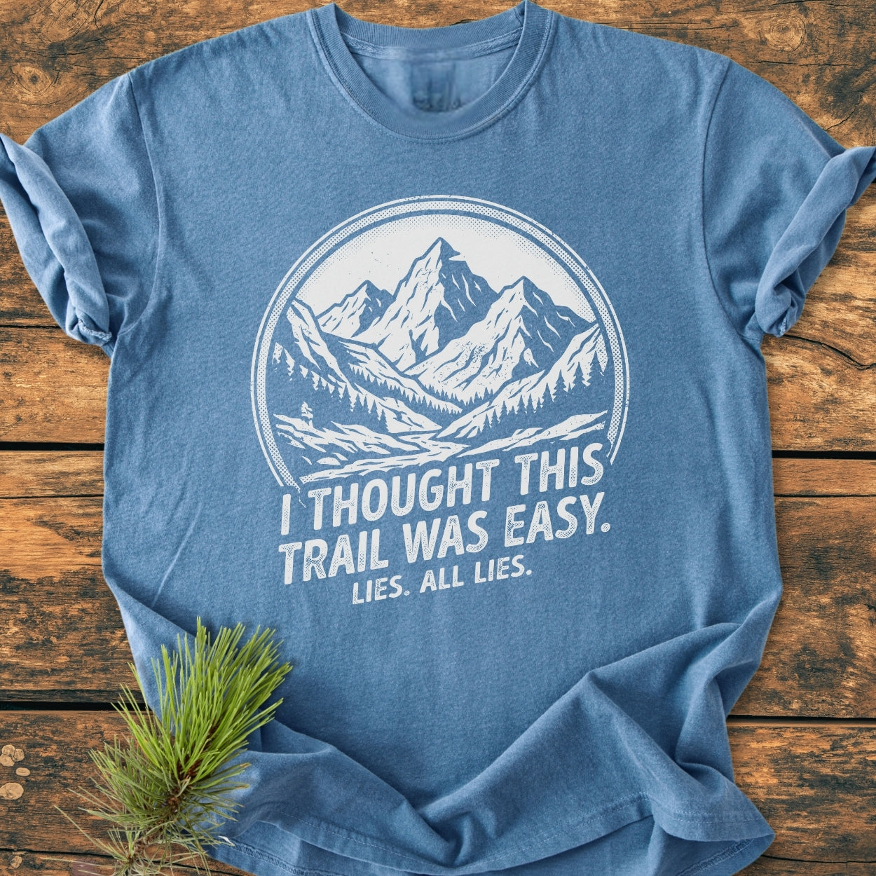 Lies, All Lies - Vintage Wash Tee: Heathered-blue crew-neck T-shirt with mountain graphic and humorous text, laid flat on a wooden surface, accented with a pine sprig.