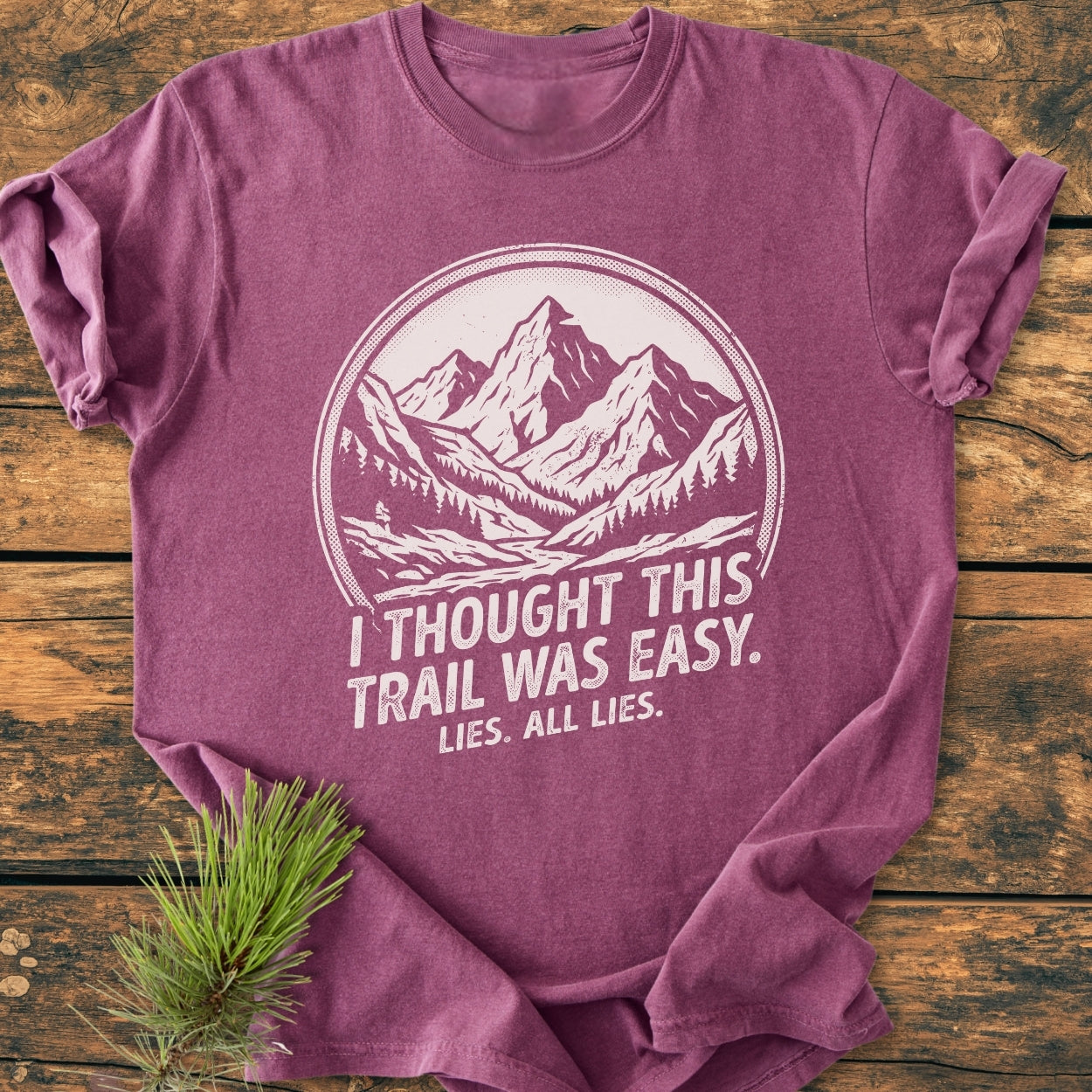 Lies, All Lies - Vintage Wash Tee featuring a mountain and pine graphic, laid flat on wood with a pine branch. Made from soft-washed, pre-shrunk 100% ring-spun cotton for comfort and durability.