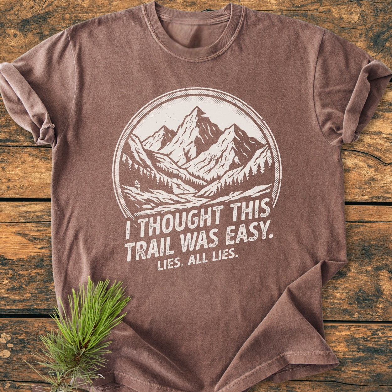Lies, All Lies - Vintage Wash Tee: Brown short-sleeved T-shirt with mountain and pine illustration, slogan printed in white. Laid on wood, accompanied by a green pine branch accent.