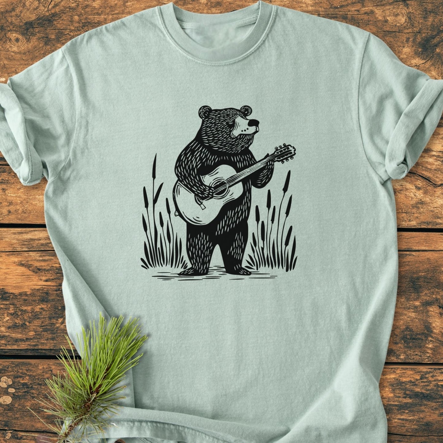Bear Playing Guitar - Vintage Wash Tee features a light-sea-foam-green hue with a black line-art bear playing guitar amid reeds, laid on a rustic wooden surface.