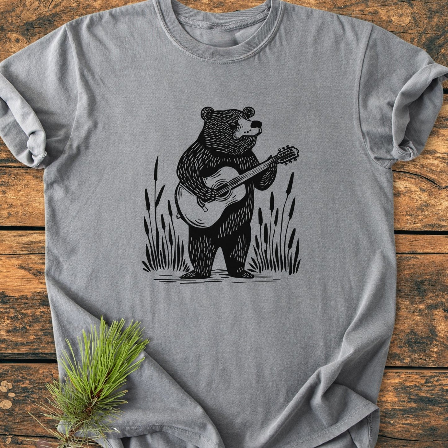 Bear Playing Guitar - Vintage Wash Tee features a flat-lay heather-gray T-shirt with a black line-art bear playing guitar, surrounded by cattails. A pine sprig accents the rustic presentation.