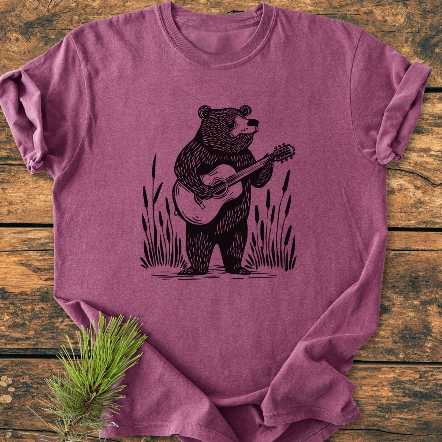 Bear Playing Guitar - Vintage Wash Tee: A soft, heathered purple T-shirt featuring a black line-drawing of a bear with a guitar, flanked by reeds, laid on a wooden surface with pine needles.