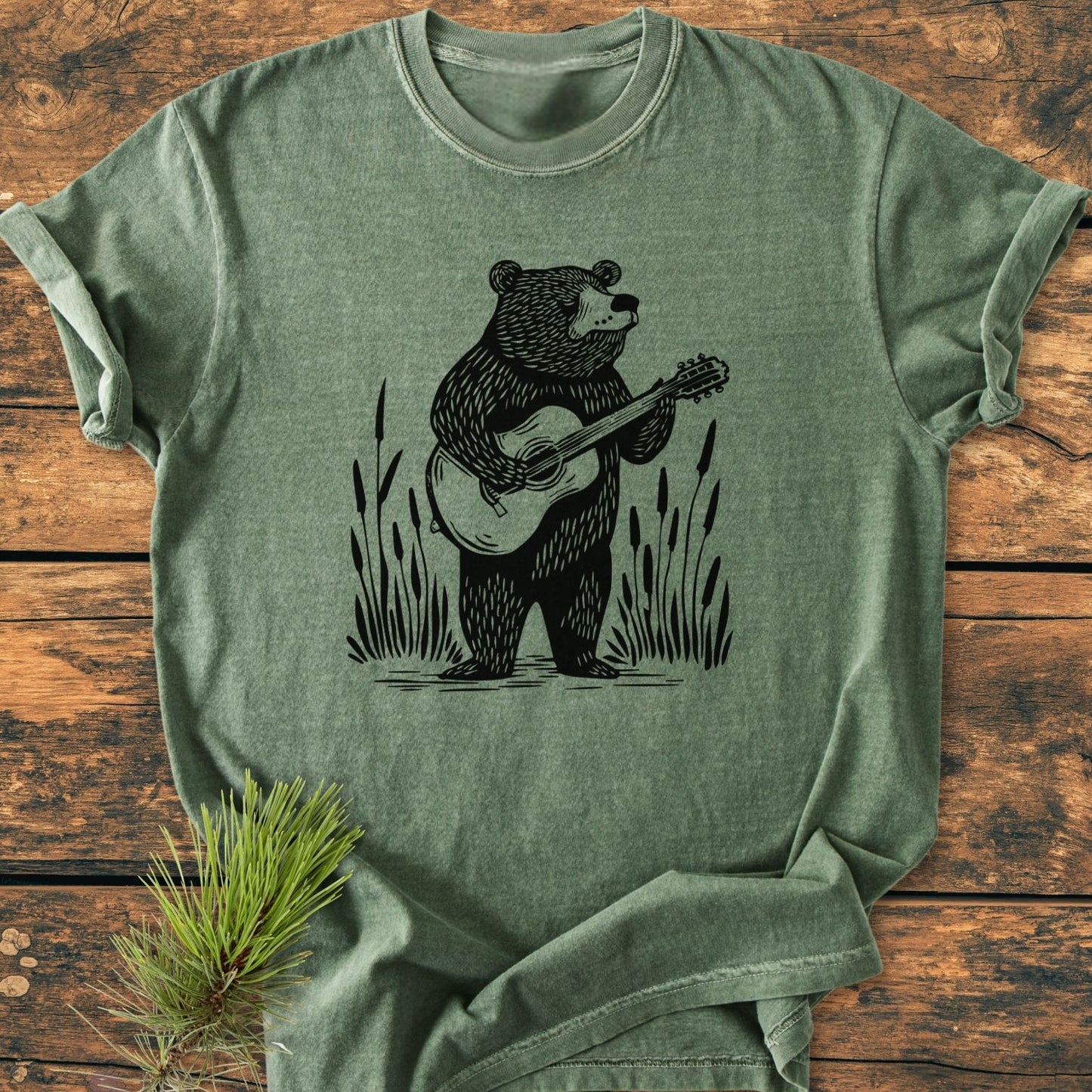 Bear Playing Guitar - Vintage Wash Tee features a stylized bear with guitar, framed by reeds. The heather-green T-shirt highlights comfort and durability, with a pine sprig accentuating its rustic charm.