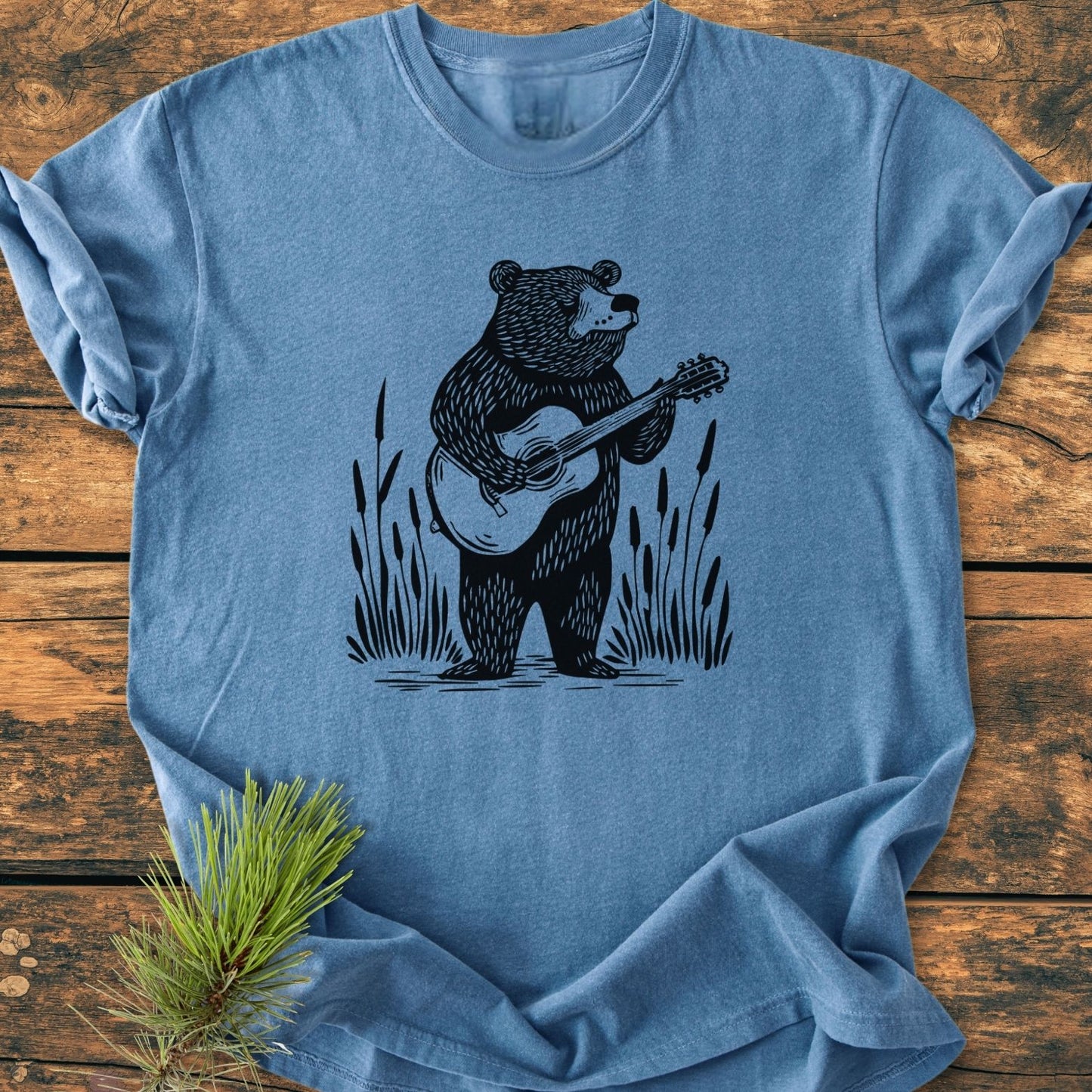Bear Playing Guitar - Vintage Wash Tee features a hand-drawn bear with guitar illustration on a heather-blue crew-neck, laid flat on wood. Made from soft, durable, pre-shrunk 100% ring-spun cotton.