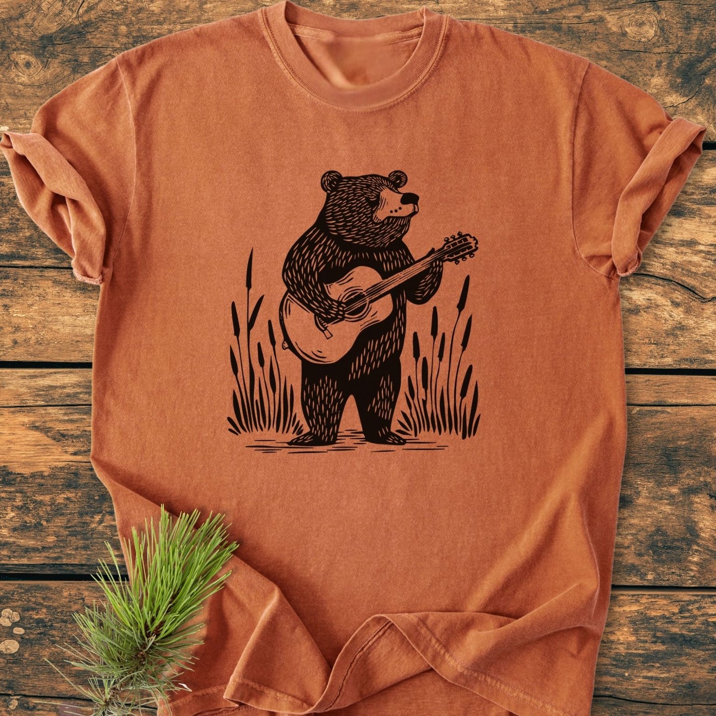 Bear Playing Guitar - Vintage Wash Tee featuring a black line-drawing of a bear with a guitar, laid on wood with pine accent, showcasing its relaxed fit and durable, soft-washed cotton fabric.