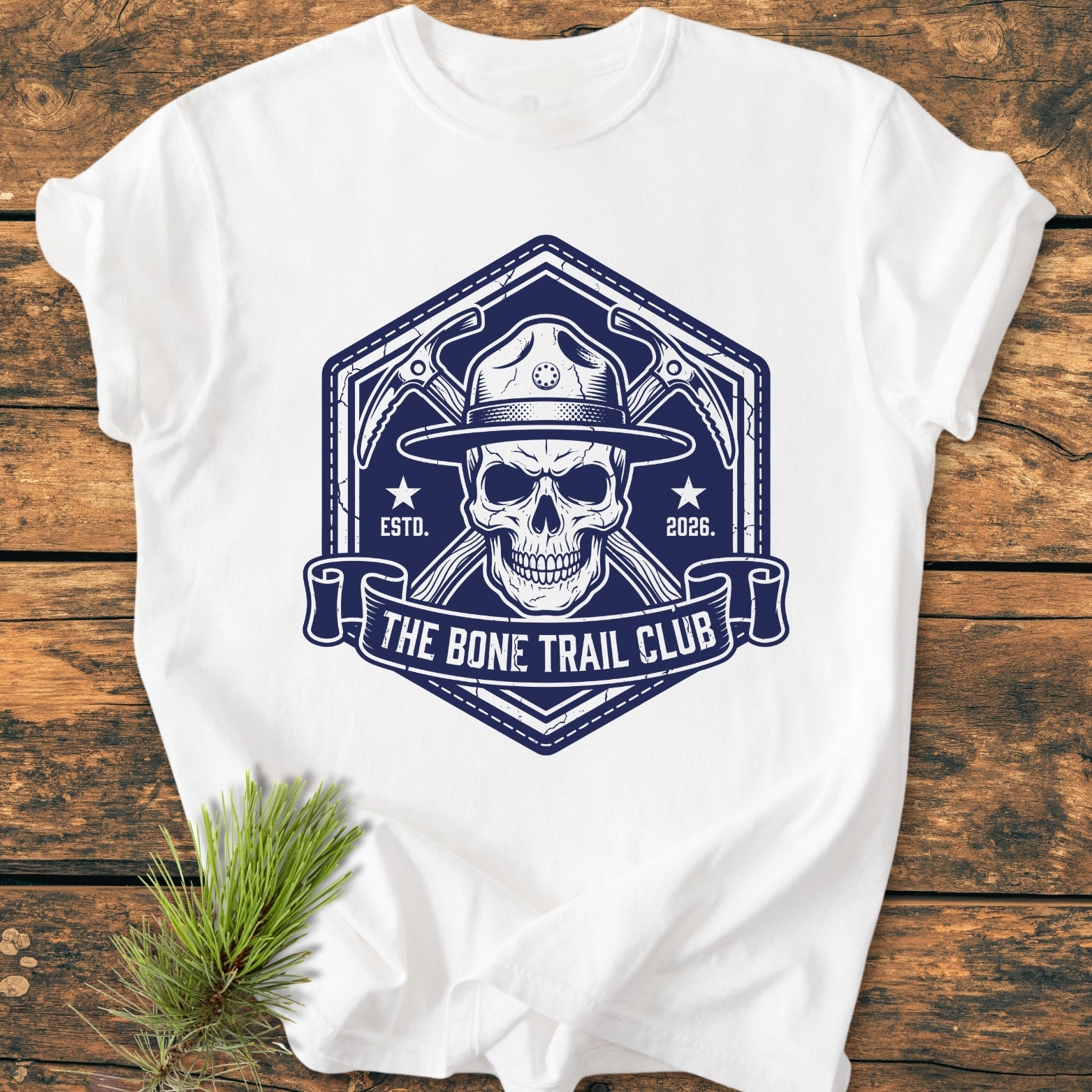 The Bone Trail Club - Vintage Wash Tee features a dark-blue badge logo with a skull in a ranger hat and crossed ice-axes, laid on a wooden surface, highlighting its rugged design.