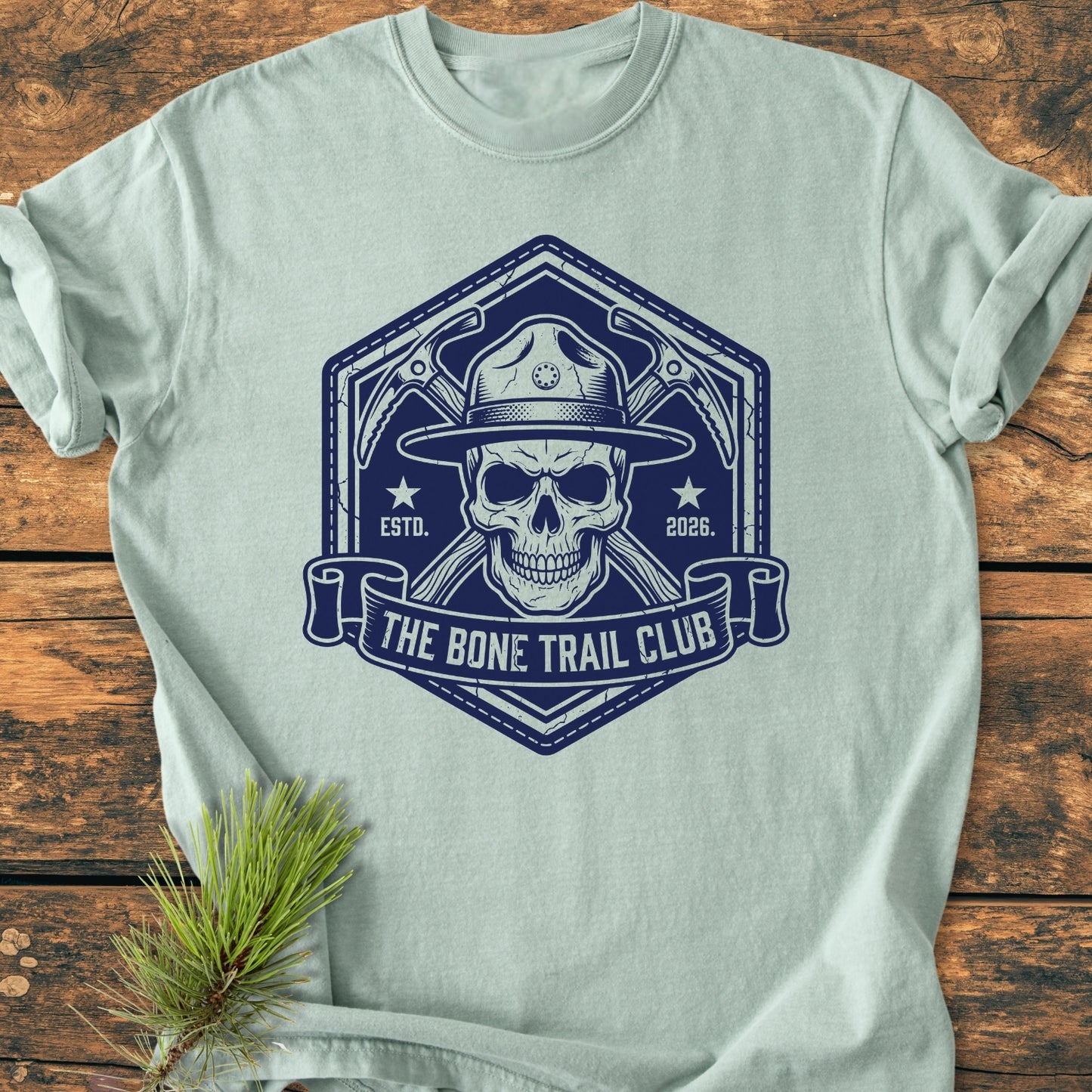 The Bone Trail Club - Vintage Wash Tee features a navy-blue emblem of a skull with ice-axes and a ranger hat, framed in a hexagonal badge, laid on a wooden surface.