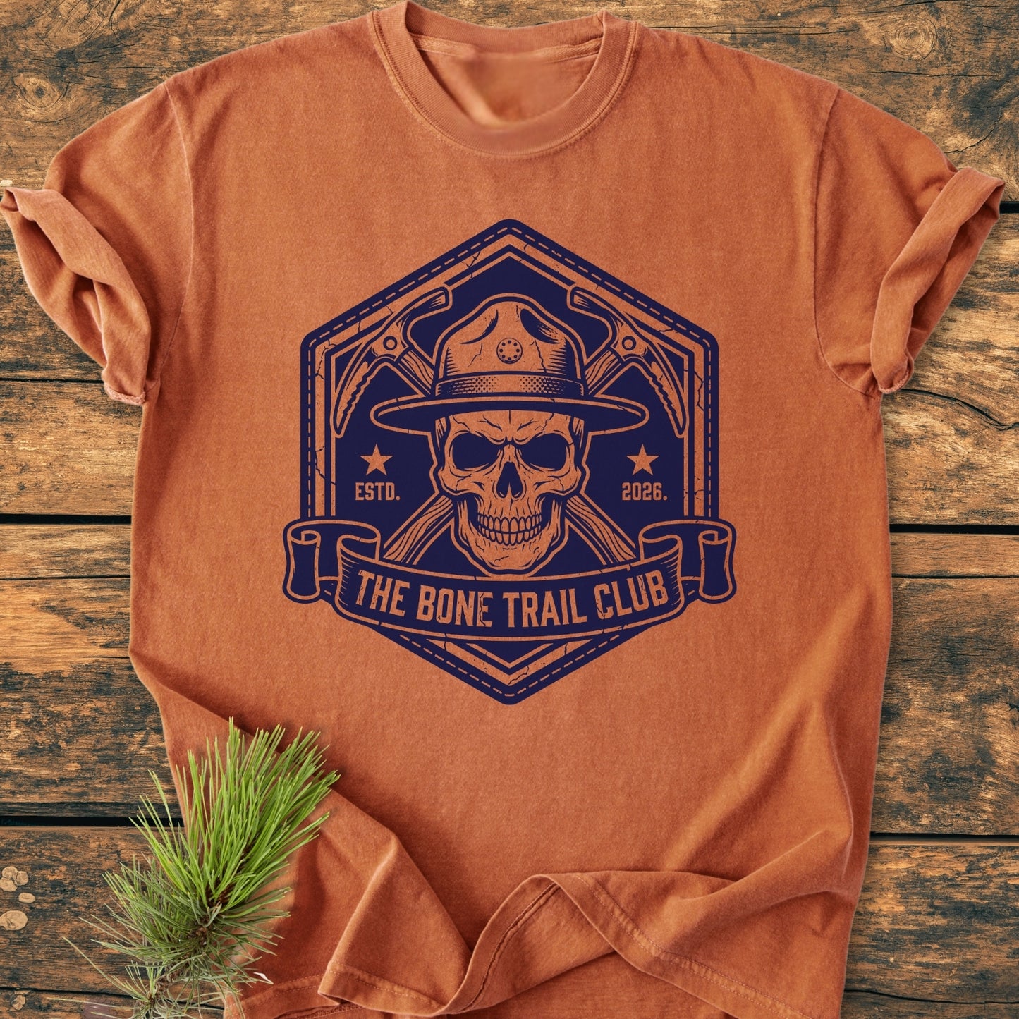 The Bone Trail Club - Vintage Wash Tee features a bold skull graphic with a ranger hat, crossed ice-axes, and a ribbon banner, laid on a wooden surface with a pine sprig adjacent.