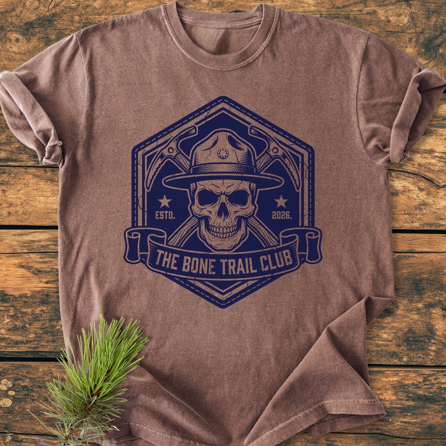 The Bone Trail Club - Vintage Wash Tee, featuring a skull with a ranger hat and crossed ice-axes design on a brown heathered shirt, laid on a rustic surface with a pine sprig.