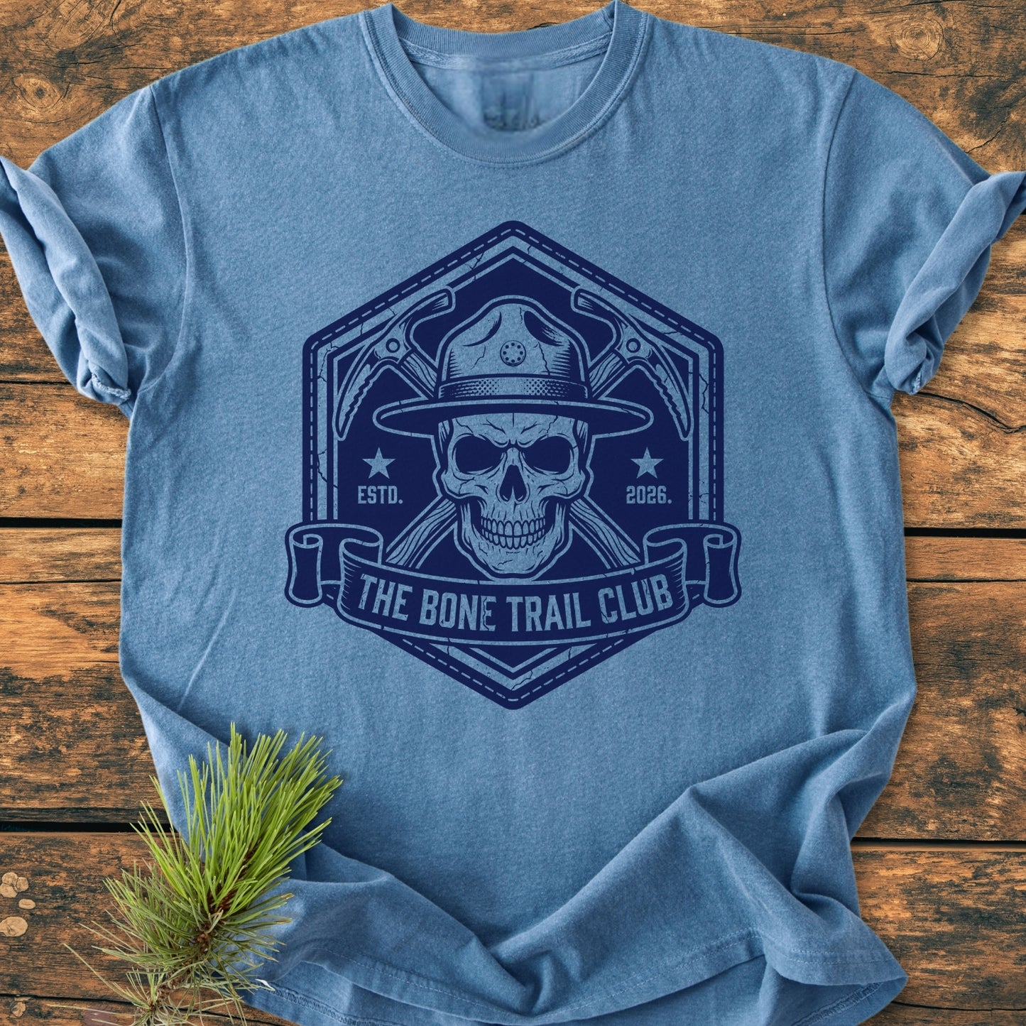 The Bone Trail Club - Vintage Wash Tee features a grinning skull with a ranger hat and crossed ice-axes, printed on a heather-blue T-shirt, laid on a wooden surface with a pine sprig.
