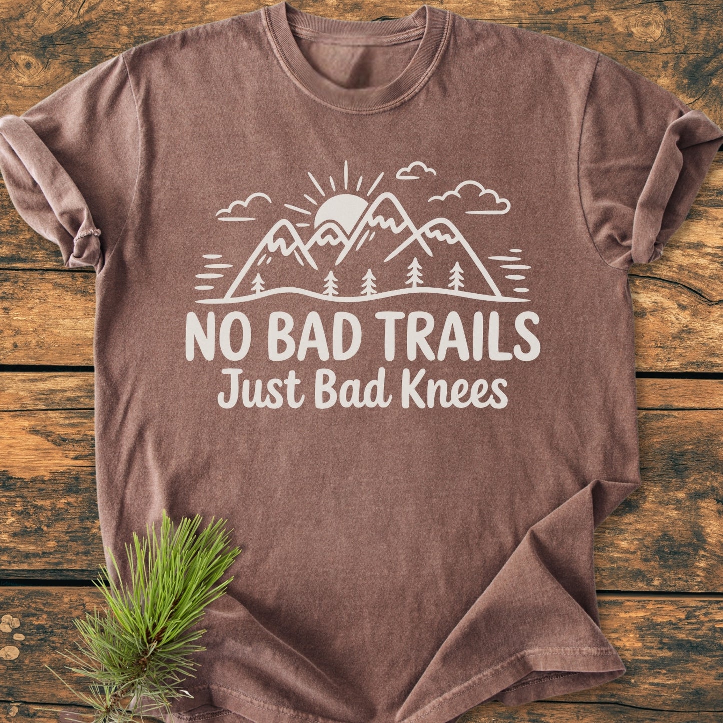 No Bad Trails - Vintage Wash Tee featuring a white graphic of mountains, trees, and sun. Laid flat, it highlights the message “NO BAD TRAILS Just Bad Knees” on a wooden surface with pine needles.