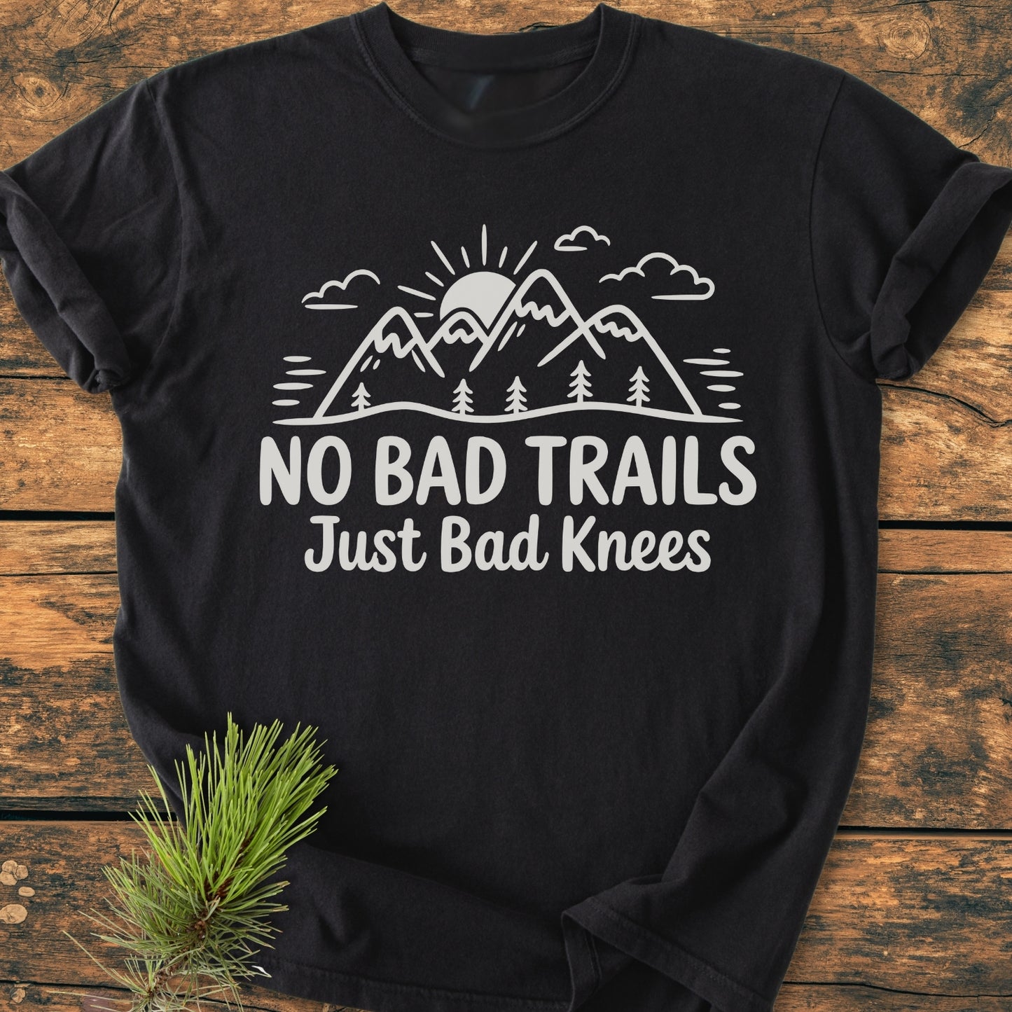 No Bad Trails - Vintage Wash Tee features a white graphic of mountains, trees, and a rising sun with the slogan “No Bad Trails Just Bad Knees,” flat-laid on wood with a pine sprig accent.