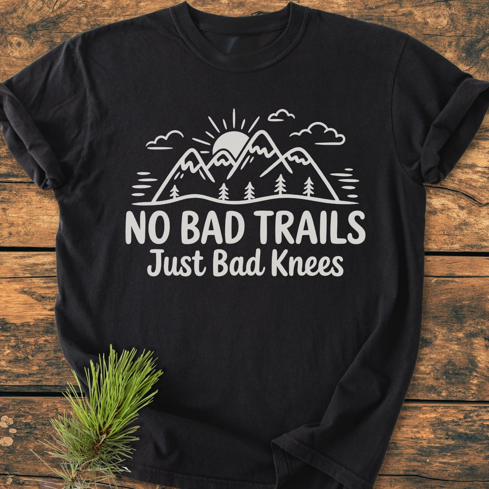 No Bad Trails - Vintage Wash Tee features a white graphic of mountains, trees, and a rising sun with the slogan “No Bad Trails Just Bad Knees,” flat-laid on wood with a pine sprig accent.