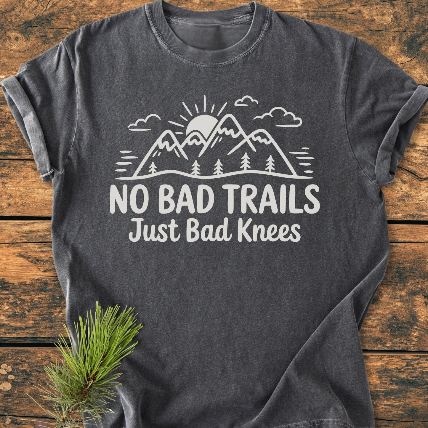 No Bad Trails - Vintage Wash Tee features a charcoal-gray design with mountains, sun, and pine trees graphic, slogan “NO BAD TRAILS Just Bad Knees,” on rustic wood, with pine branch accent.
