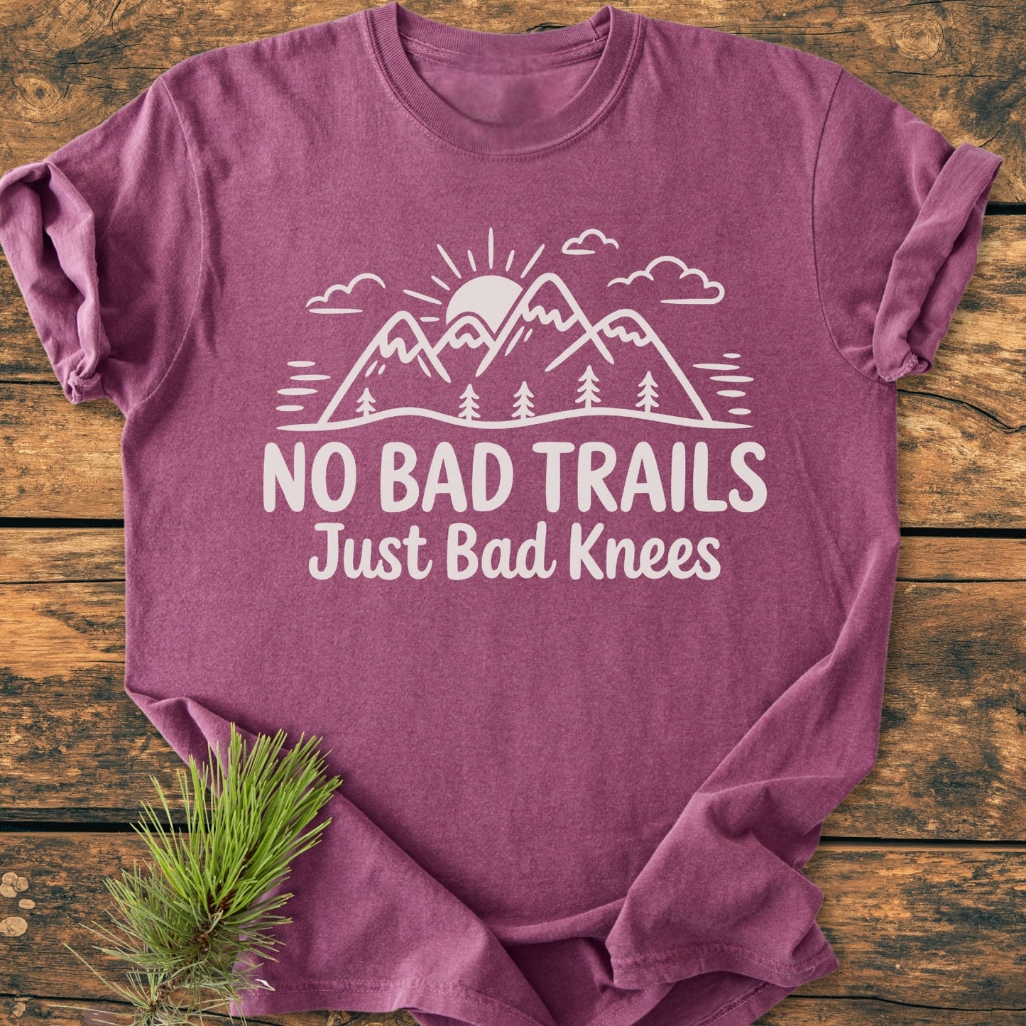 No Bad Trails - Vintage Wash Tee features a white graphic of mountains and trees on a plum t-shirt, laid flat with a pine branch accent, showcasing a relaxed, cozy fit.