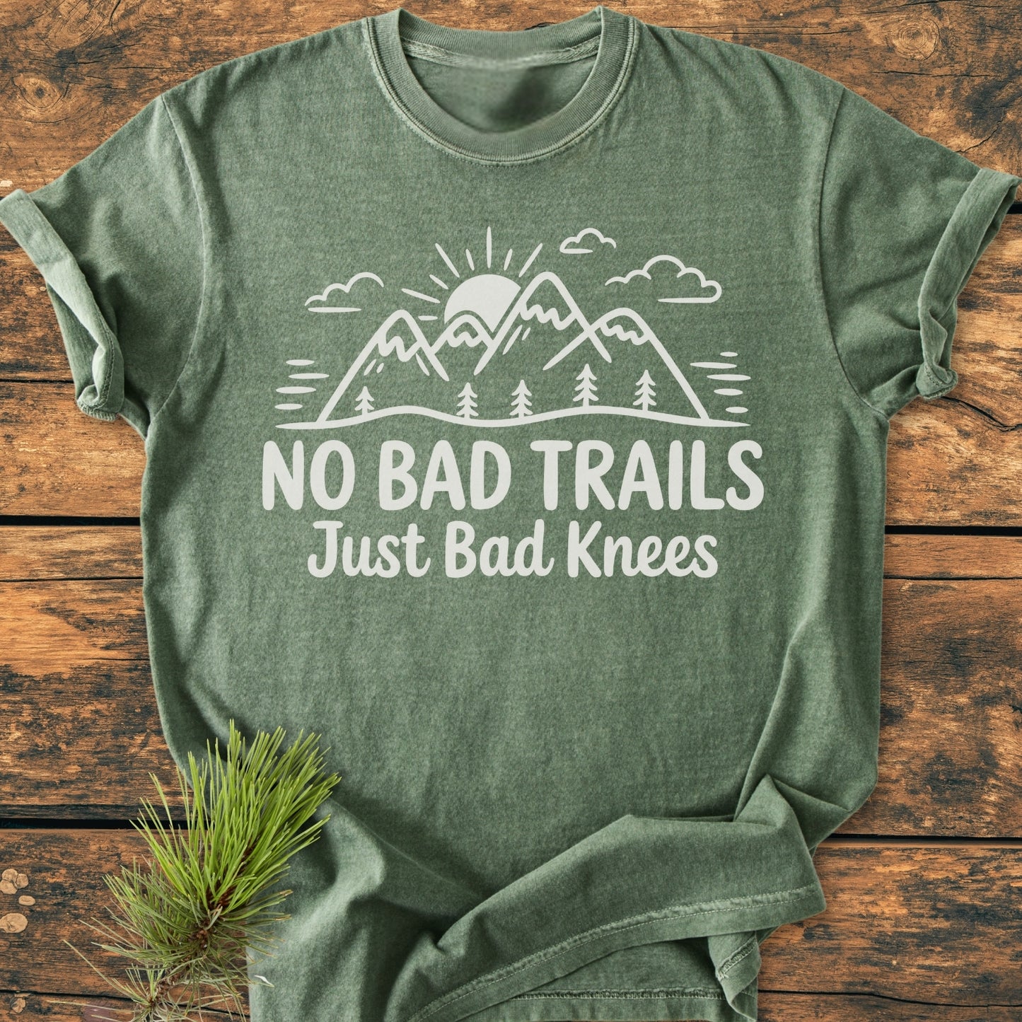 No Bad Trails - Vintage Wash Tee featuring a white mountain and sun design on heather-green fabric, laid on wood. Includes pine sprig accent, highlighting the tee's outdoor-themed slogan and rustic appeal.