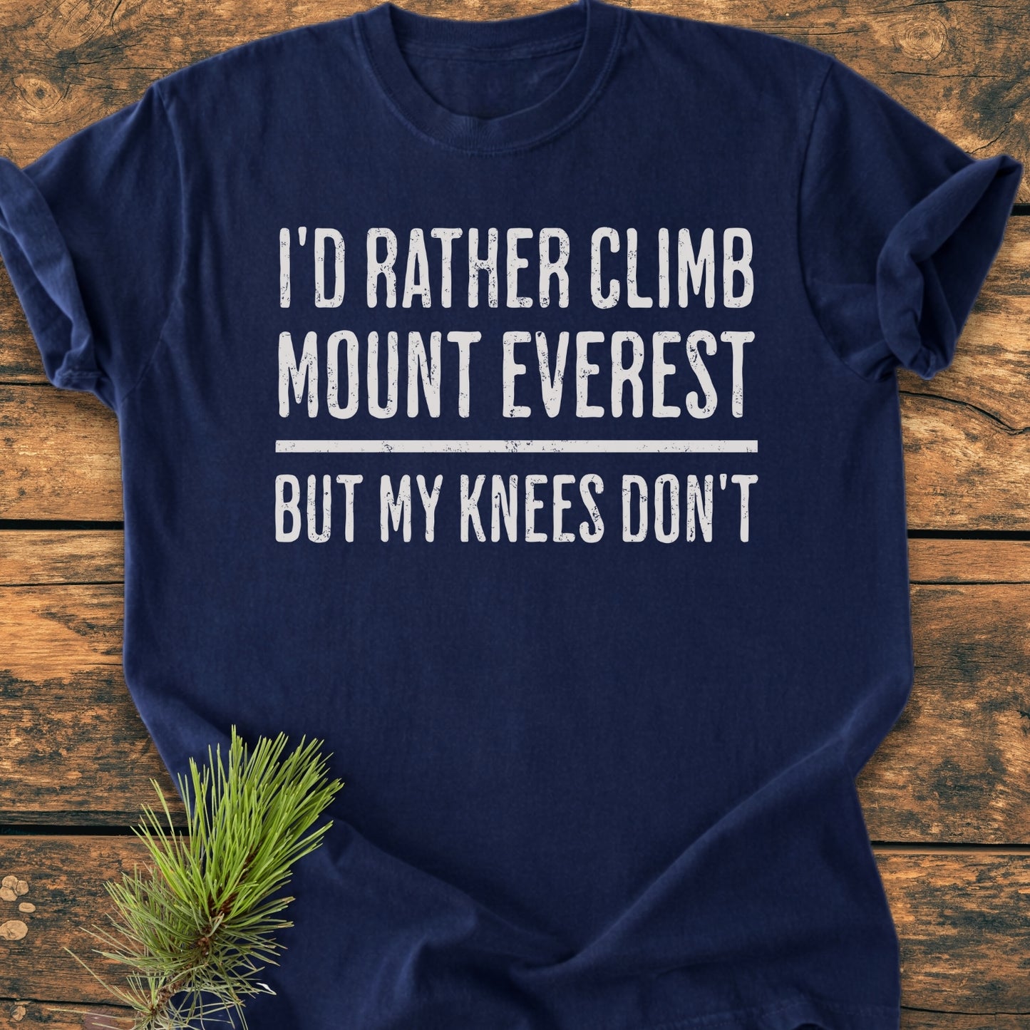 “I’d Rather Climb Mount Everest - Vintage Wash Tee” in navy-blue, laid on wood with distressed white slogan, showcasing its relaxed fit and soft, garment-dyed cotton texture. Pine sprig decorates the corner.