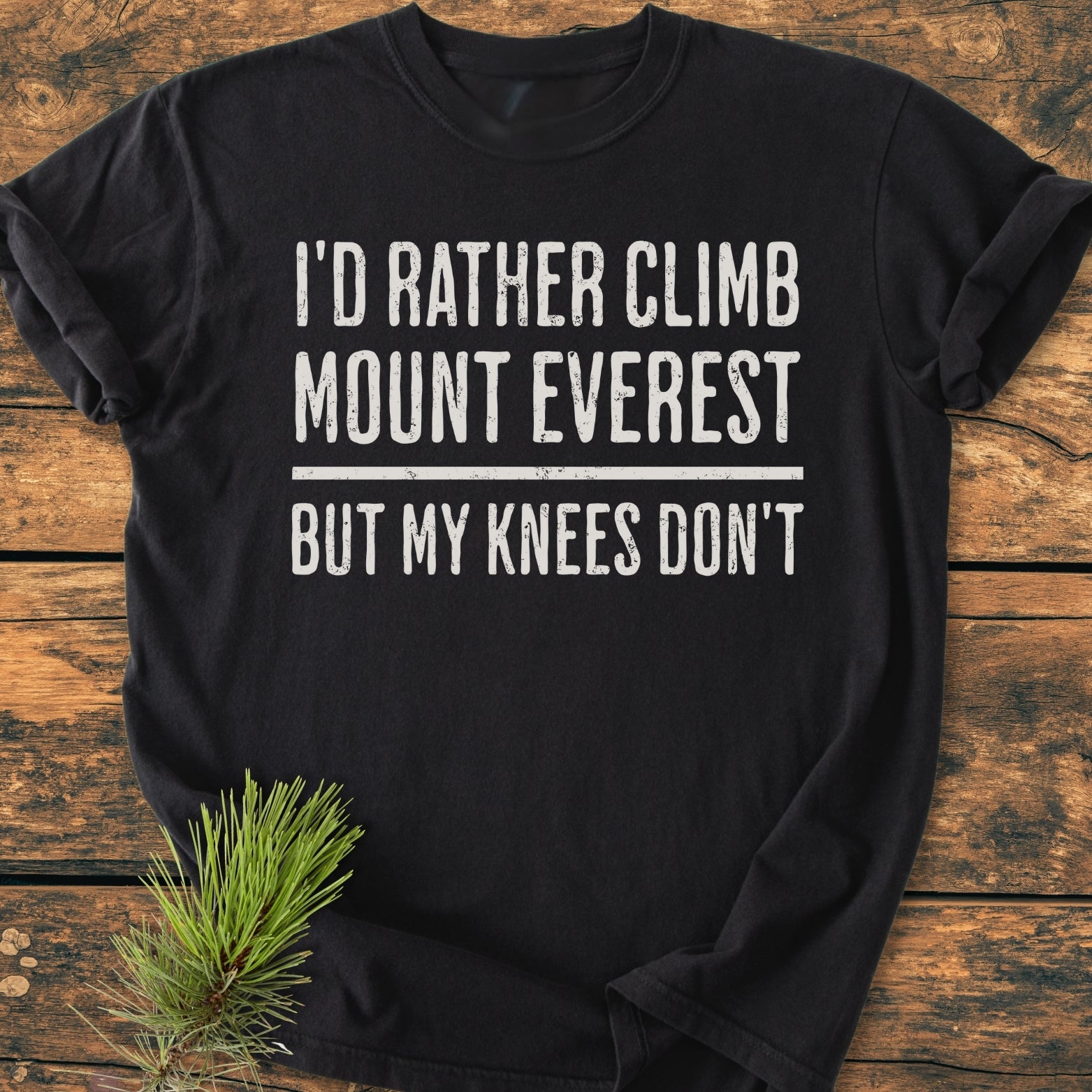 I'd Rather Climb Mount Everest - Vintage Wash Tee laid flat on wood, featuring distressed white lettering. A small pine sprig accents the scene, highlighting the shirt's comfort and relaxed fit.