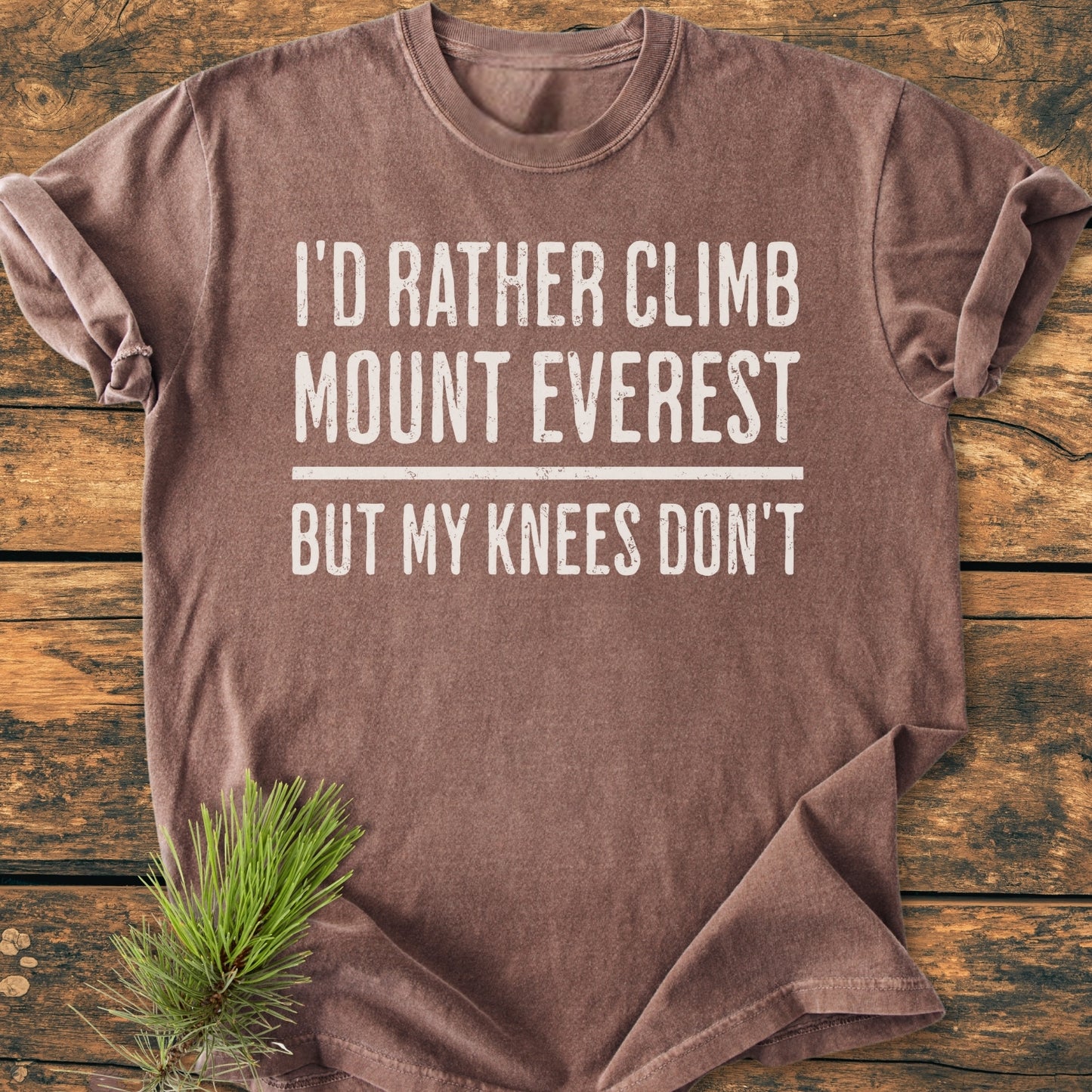 I'd Rather Climb Mount Everest - Vintage Wash Tee features bold white text on a brown short-sleeved shirt, laid on rustic wood with pine needles accentuating its cozy, relaxed fit.
