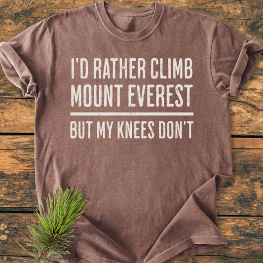 I'd Rather Climb Mount Everest - Vintage Wash Tee features bold white text on a brown short-sleeved shirt, laid on rustic wood with pine needles accentuating its cozy, relaxed fit.