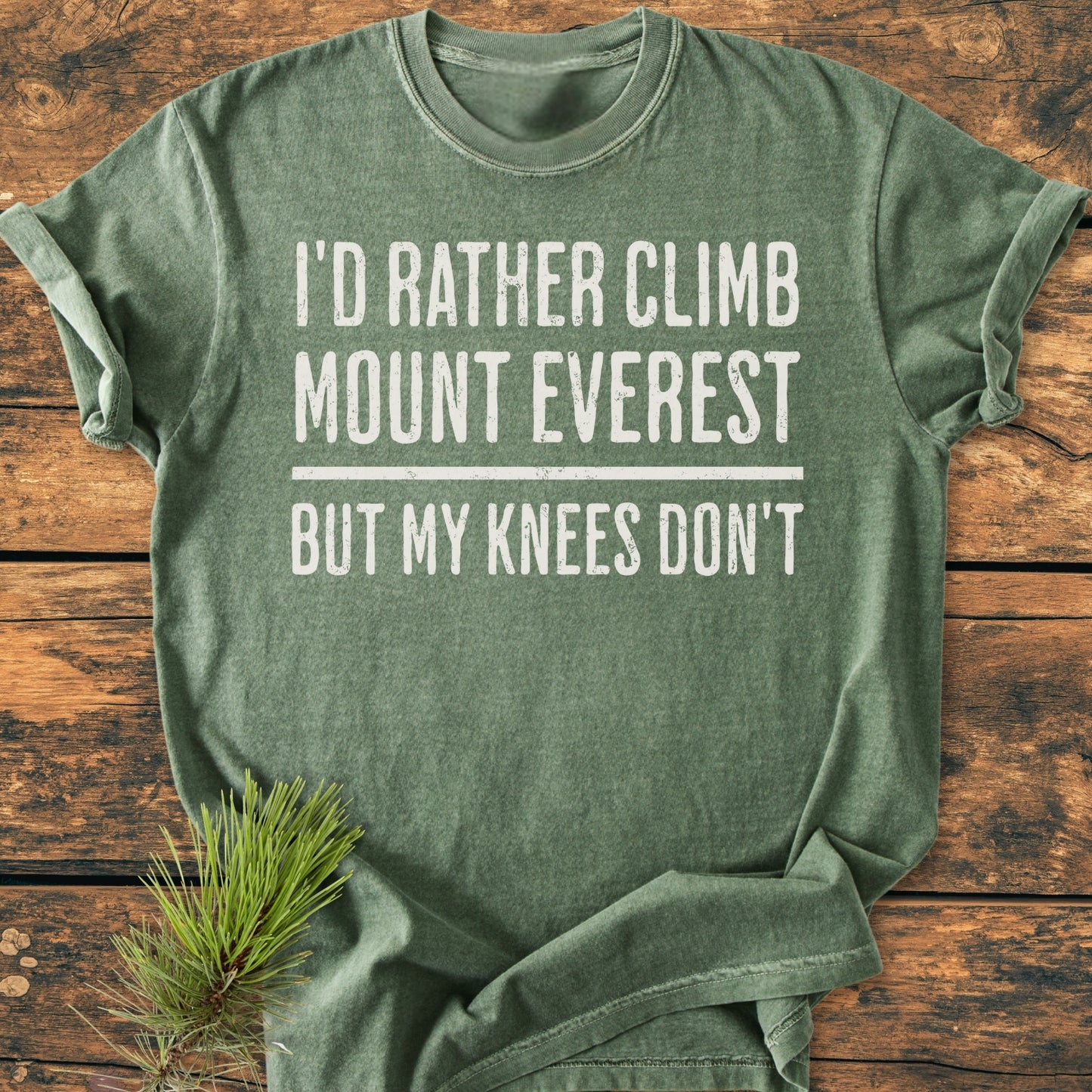 Heather-green I'd Rather Climb Mount Everest - Vintage Wash Tee laid flat on wood surface, featuring bold white lettering, styled with a pine sprig. Relaxed fit, soft-washed, ring-spun cotton.