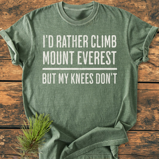 Heather-green I'd Rather Climb Mount Everest - Vintage Wash Tee laid flat on wood surface, featuring bold white lettering, styled with a pine sprig. Relaxed fit, soft-washed, ring-spun cotton.