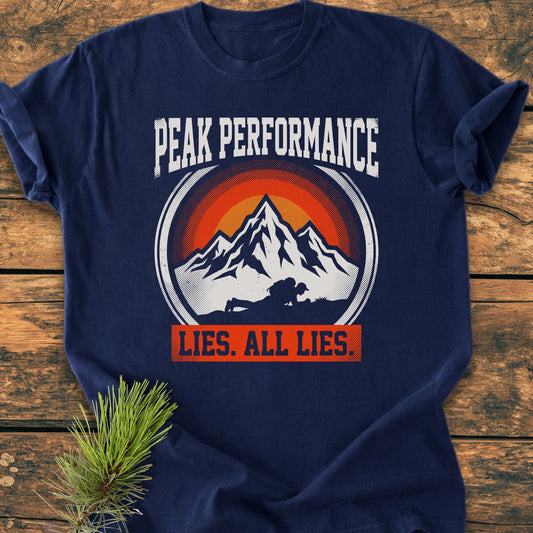 Peak Performance - Vintage Wash Tee, featuring a circular mountain peak graphic with a sunset backdrop and bold text, laid flat on a wooden surface with a decorative pine sprig beside it.