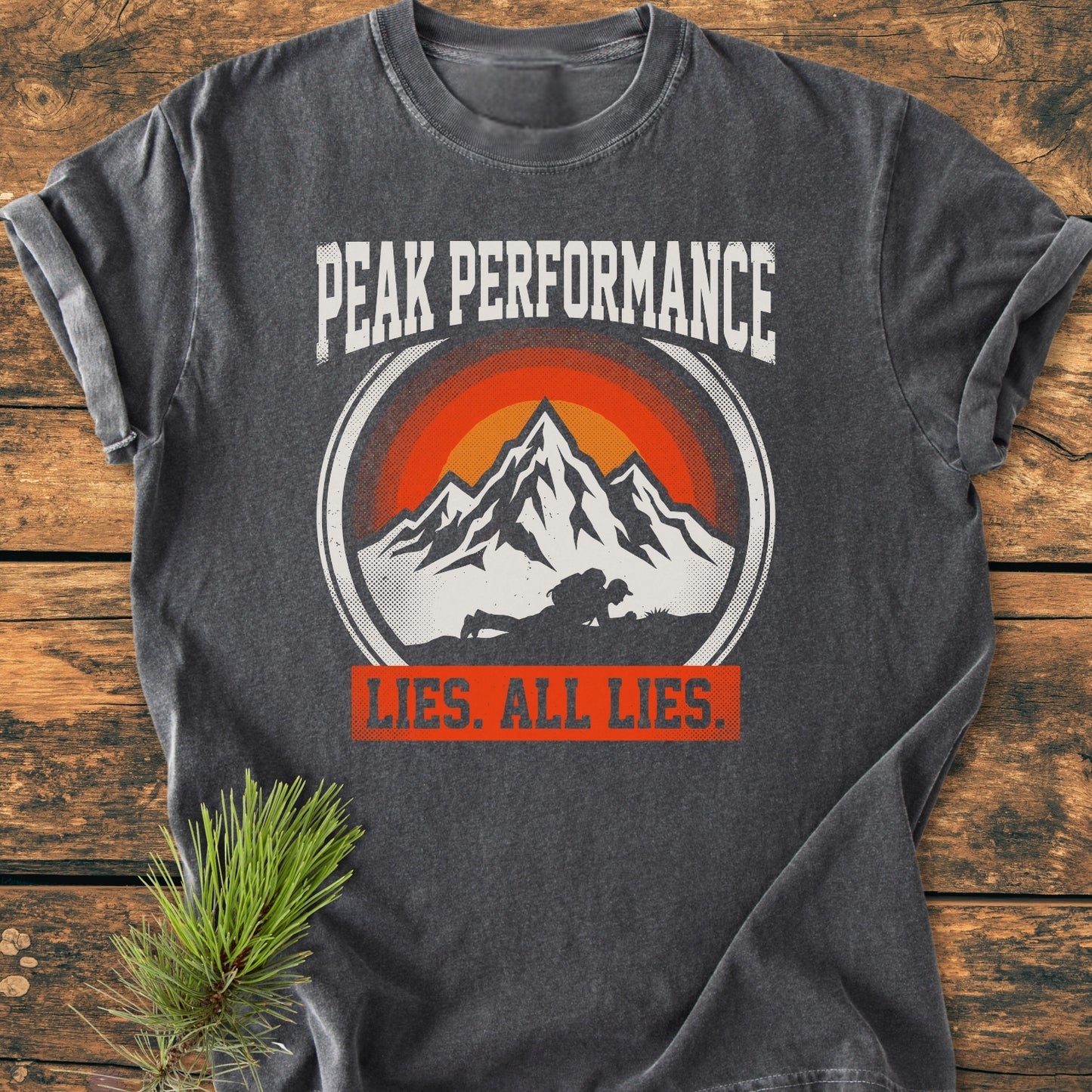 Charcoal-gray Peak Performance - Vintage Wash Tee featuring bold white text, mountain graphic with climber, and LIES. ALL LIES. banner, laid on wood with pine sprig accent.