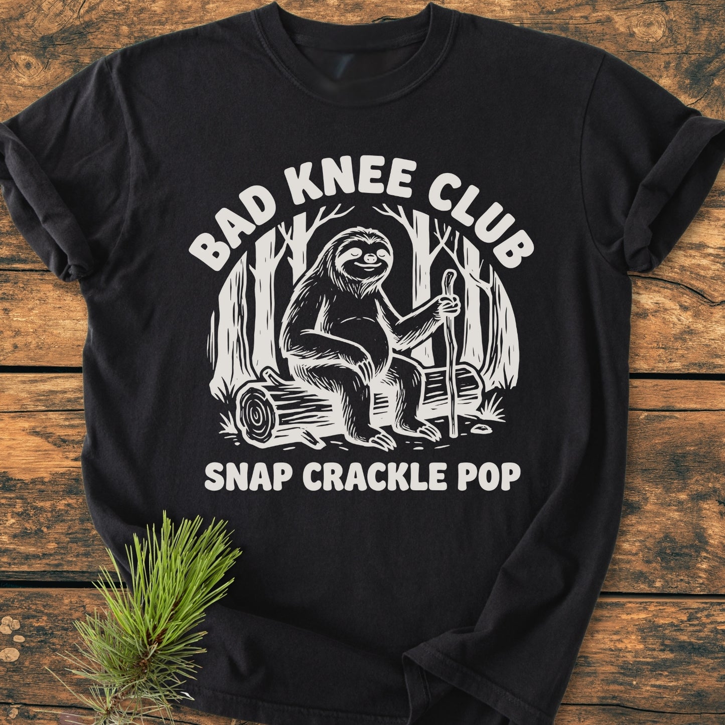 Bad Knee Club - Vintage Wash Tee features a smiling sloth with a walking stick, set in a forest scene on a soft, garment-dyed black T-shirt, styled with a spruce sprig.