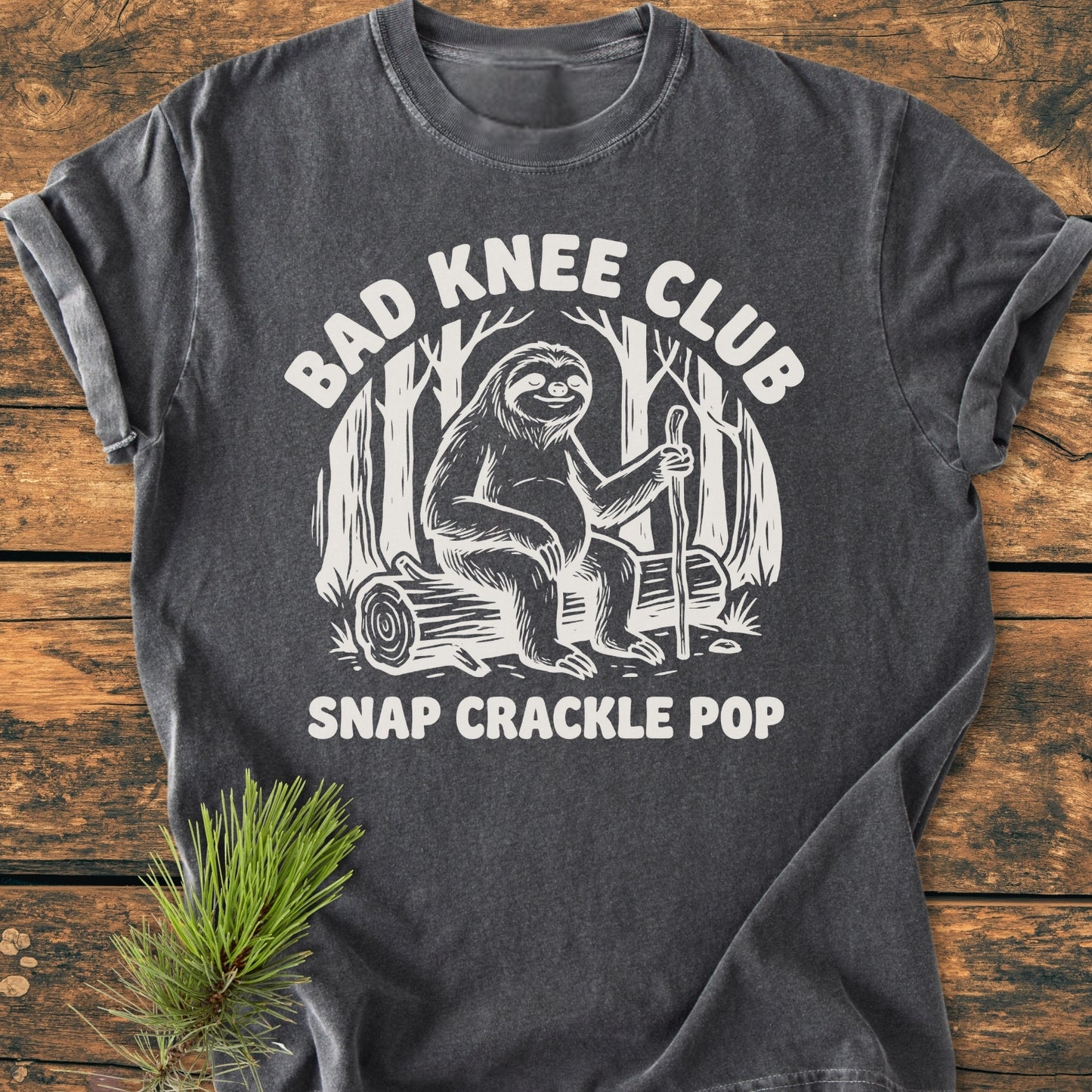 Bad Knee Club - Vintage Wash Tee features a white sloth graphic with text on a charcoal-gray T-shirt, laid flat beside a pine branch, emphasizing a cozy, relaxed fit and durable design.