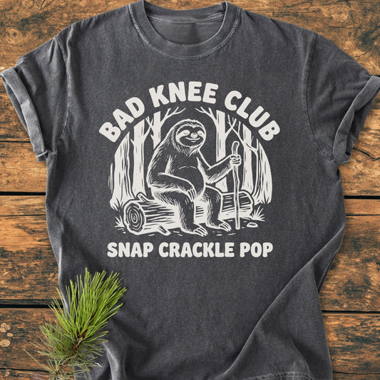 Bad Knee Club - Vintage Wash Tee features a white sloth graphic with text on a charcoal-gray T-shirt, laid flat beside a pine branch, emphasizing a cozy, relaxed fit and durable design.