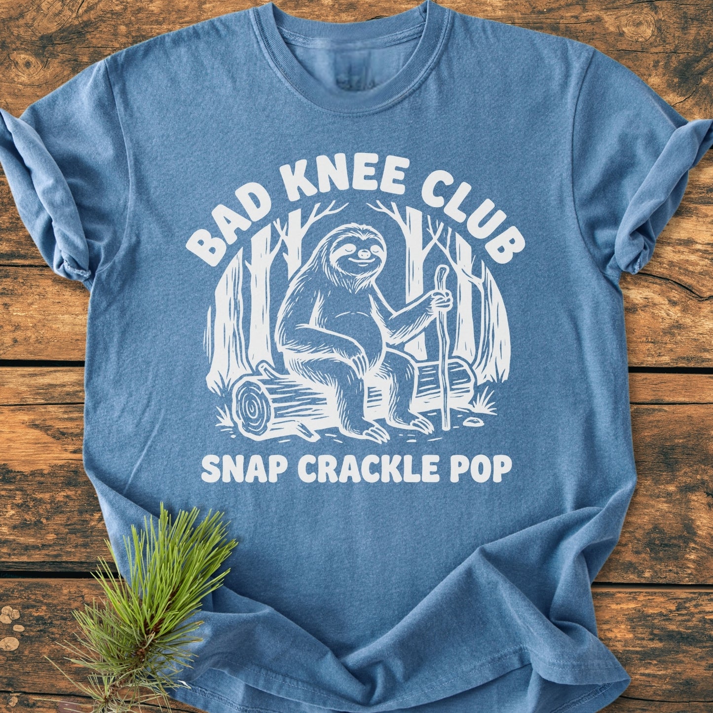 Bad Knee Club - Vintage Wash Tee featuring a cartoon sloth with a walking stick, printed on a heather-blue, soft-washed, garment-dyed shirt, styled with a pine sprig on rustic wood.