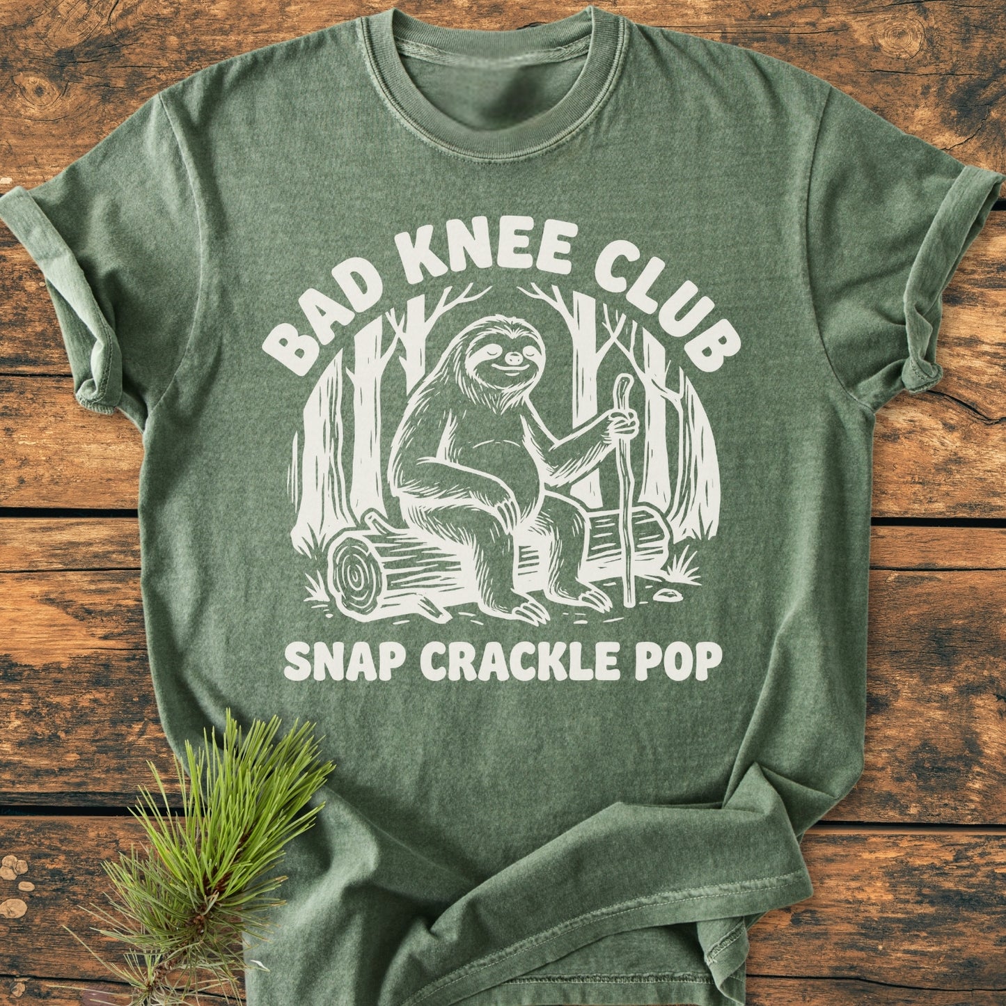 Bad Knee Club - Vintage Wash Tee features a cartoon sloth with a walking stick in a woodland scene, screen-printed on a heather-green T-shirt, styled with a pine sprig.