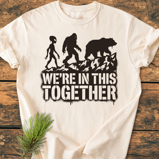 In This Together - Vintage Wash Tee features black graphics of an alien, Bigfoot, and bear on rocky ridges with a slogan. Laid on a wooden surface, it’s cozy and durable.