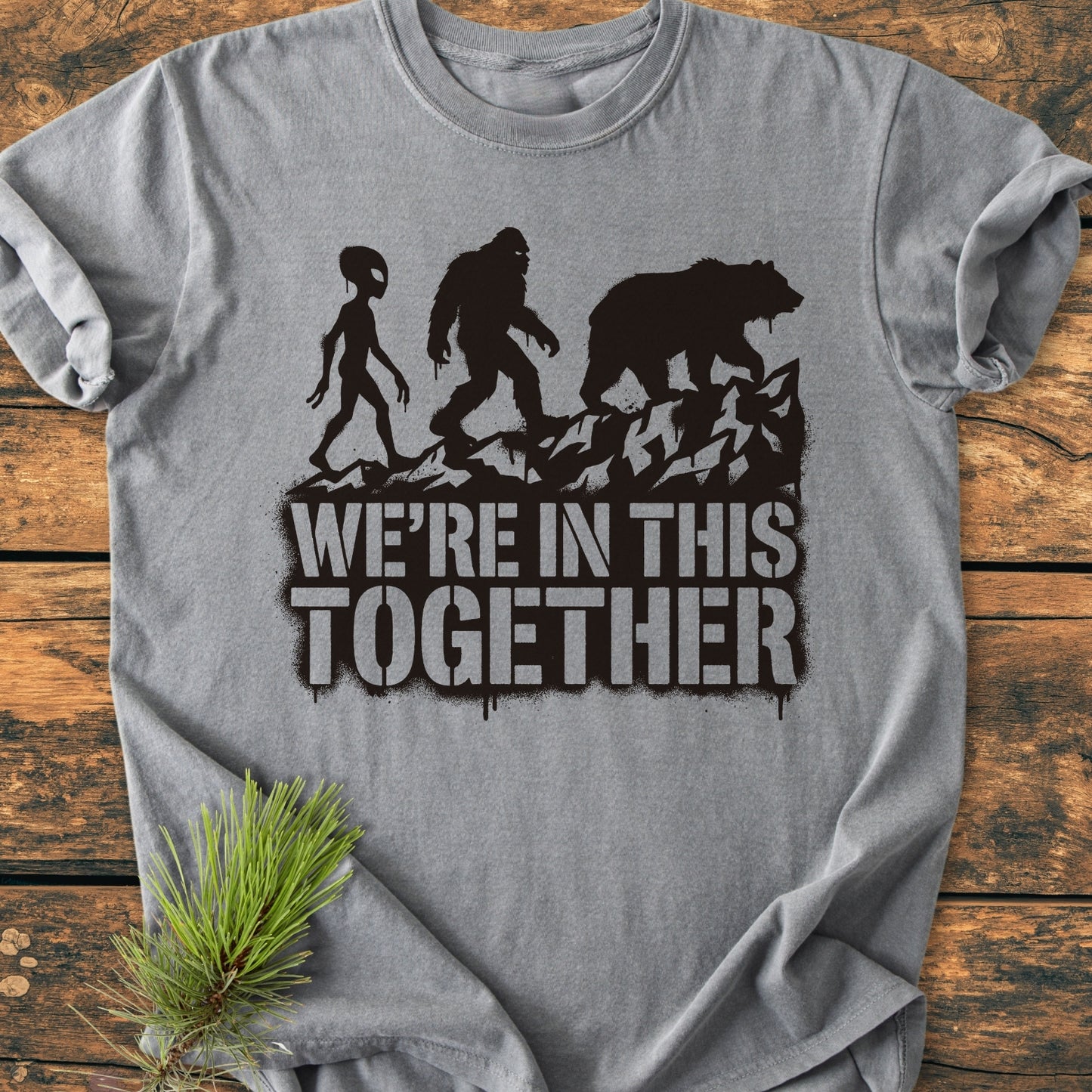 In This Together - Vintage Wash Tee features a heather-gray design with alien, Bigfoot, and bear silhouettes and WE'RE IN THIS TOGETHER text, styled with a pine sprig on a wooden surface.