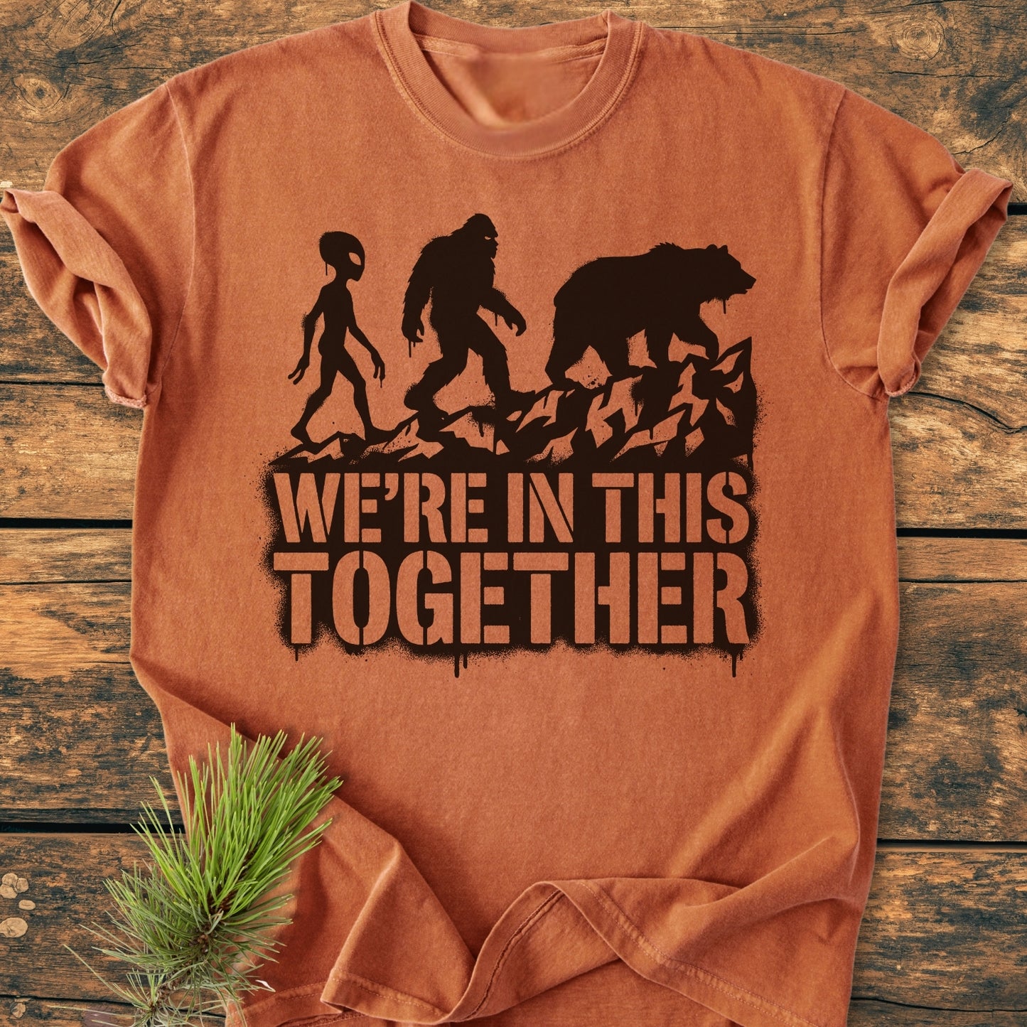 In This Together - Vintage Wash Tee features black silhouettes of an alien, Bigfoot, and bear with the slogan “WE’RE IN THIS TOGETHER” on an orange-rust garment laid on wood, complemented by a pine sprig.