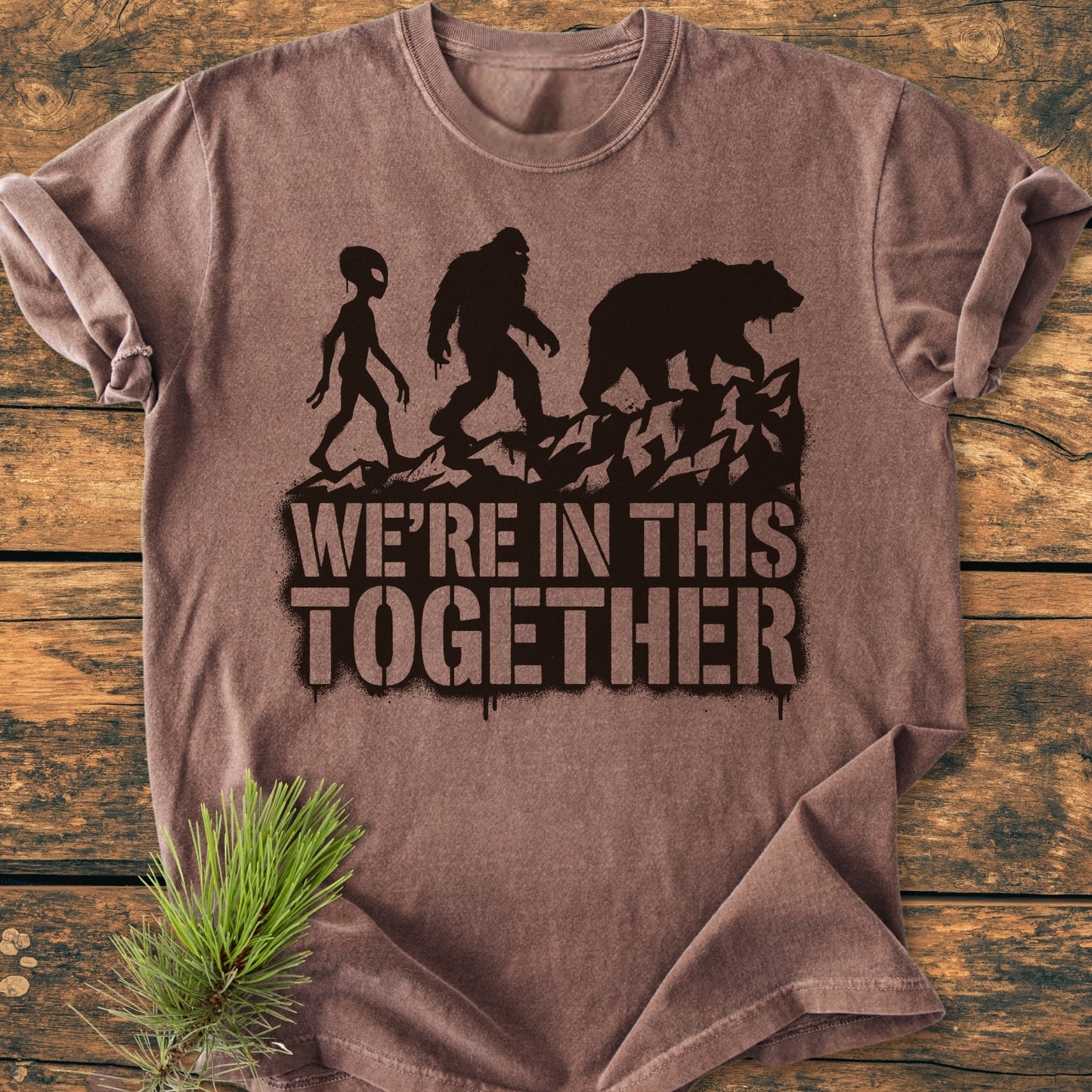 In This Together - Vintage Wash Tee: Brown t-shirt with alien, Bigfoot, and bear silhouettes, WE'RE IN THIS TOGETHER text, laid on a rustic surface with pine sprig beside.